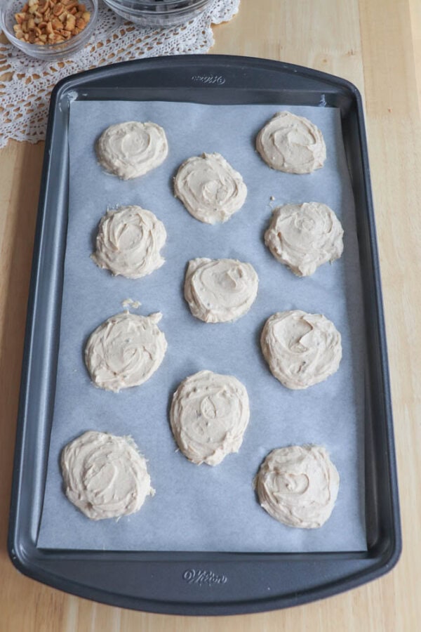 A baking tray with peanut butter yogurt portions placed on parchment paper, ready to be baked. The tray is on a wooden surface with a lace doily partially visible at the top.