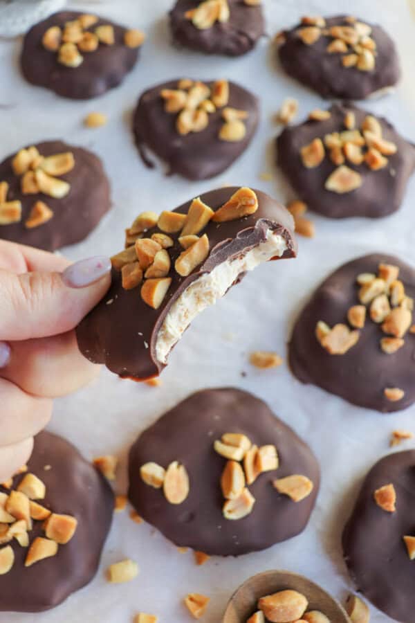 Close-up of a hand holding a chocolate-covered treat with a bite taken, revealing a nougat filling. The treat is topped with chopped nuts. More similar treats are on parchment paper in the background.