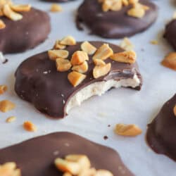 Close-up of chocolate-covered treats topped with chopped nuts, placed on parchment paper. One treat has a bite taken out, revealing a creamy filling. Scattered nut pieces add texture to the scene.