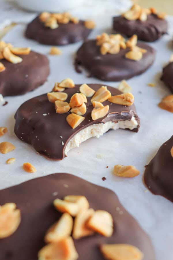 Close-up of chocolate-covered treats topped with chopped nuts, placed on parchment paper. One treat has a bite taken out, revealing a creamy filling. Scattered nut pieces add texture to the scene.