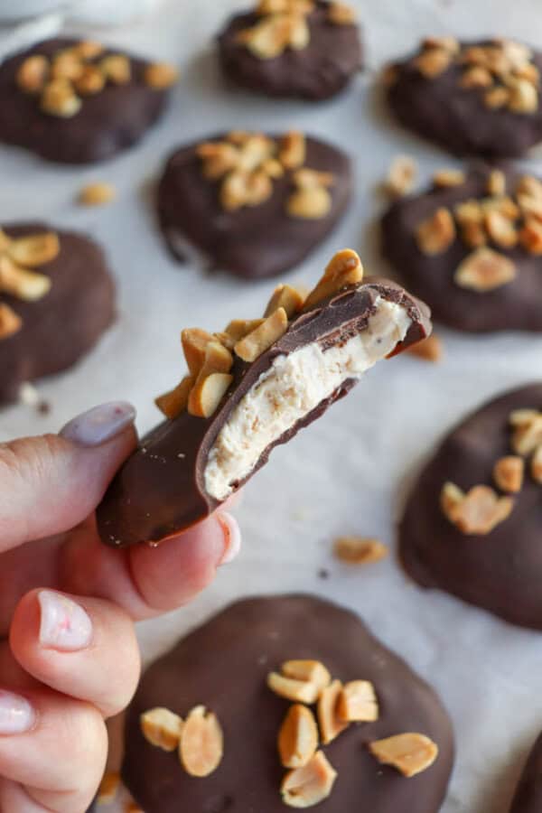 A close-up of a hand holding a chocolate-covered dessert with a bite taken out, revealing a creamy filling. The top is topped with chopped peanuts. Several similar treats are on a parchment-lined surface in the background.