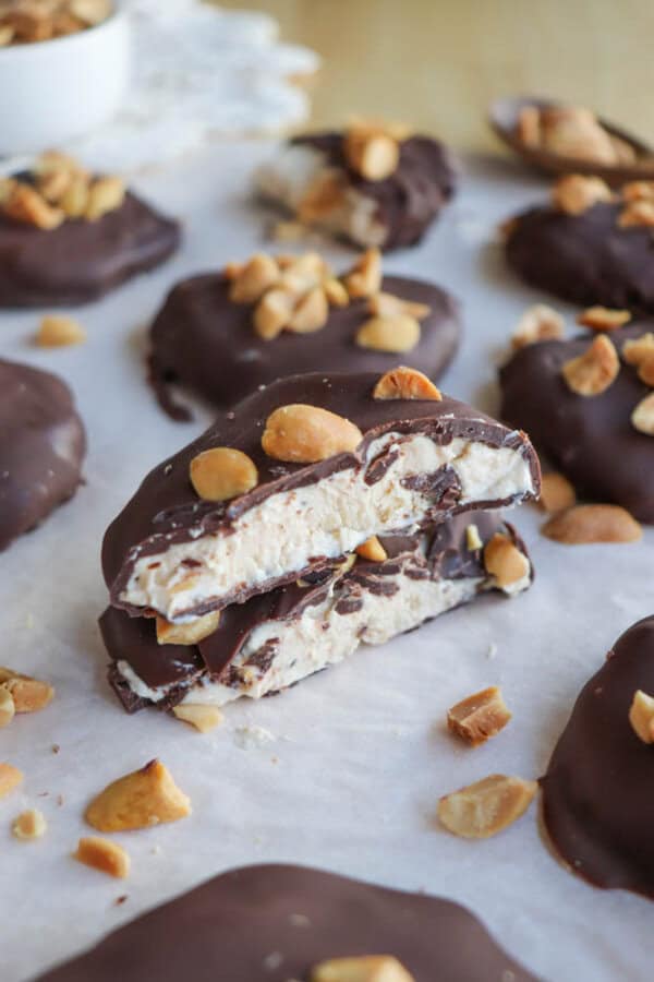 Chocolate-covered treats with a creamy nougat center are topped with chopped nuts, displayed on parchment paper. A few nuts are scattered around. One treat is broken in half, revealing the inside texture.