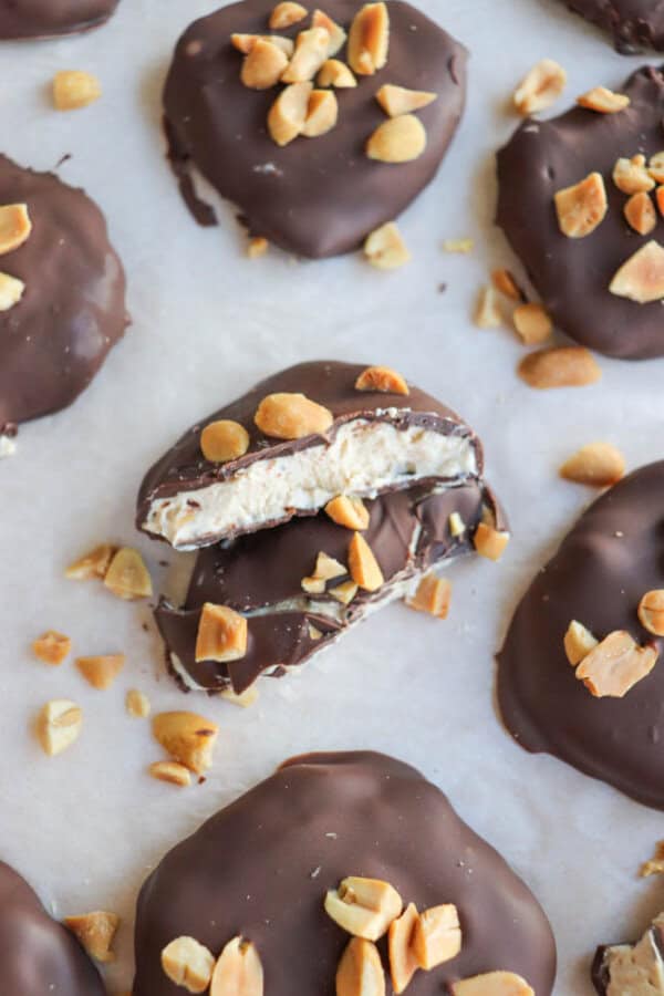 Chocolate-covered treats topped with chopped peanuts are arranged on a white surface. One treat is cut in half, revealing a creamy filling. Pieces of peanut are scattered around.