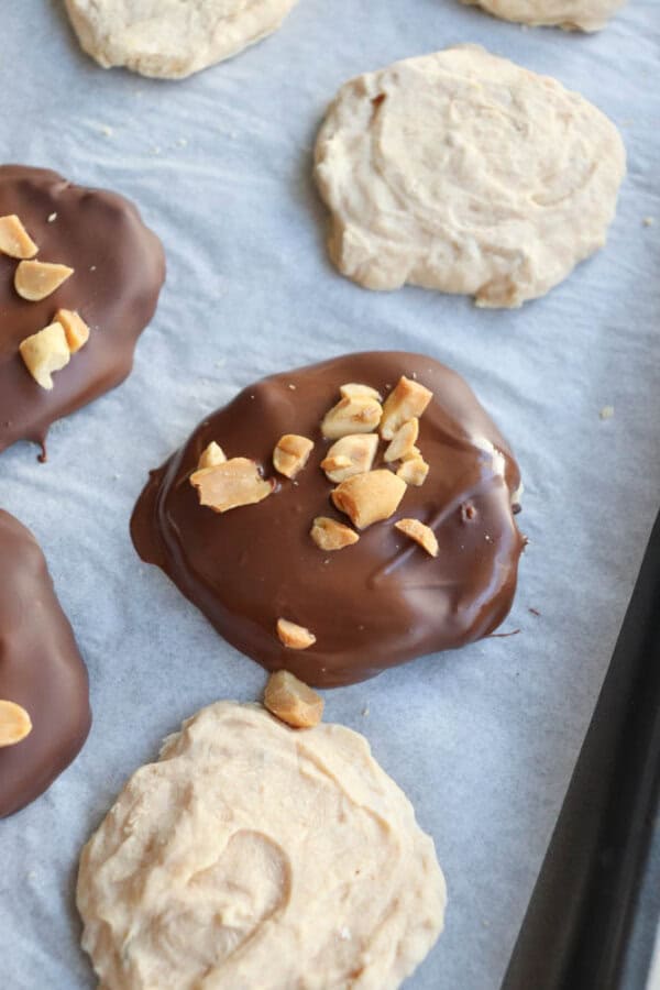 Cookies on a baking tray, some partially covered in chocolate with chopped nuts on top, and others plain. The tray is lined with parchment paper.