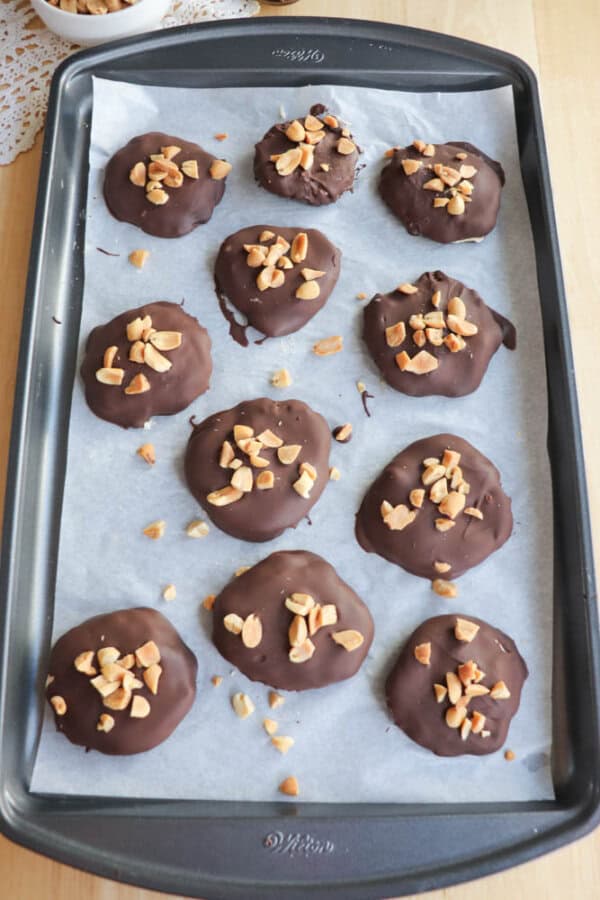 A baking tray lined with parchment paper holds 12 chocolate-coated round cookies topped with chopped nuts. The tray rests on a light wooden surface.
