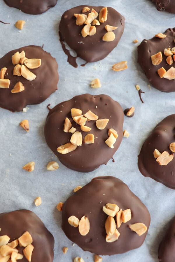 Chocolate-coated treats topped with chopped peanuts are arranged on parchment paper. Each treat is round and has a glossy, dark chocolate layer, topped with a cluster of small, golden brown peanut pieces.