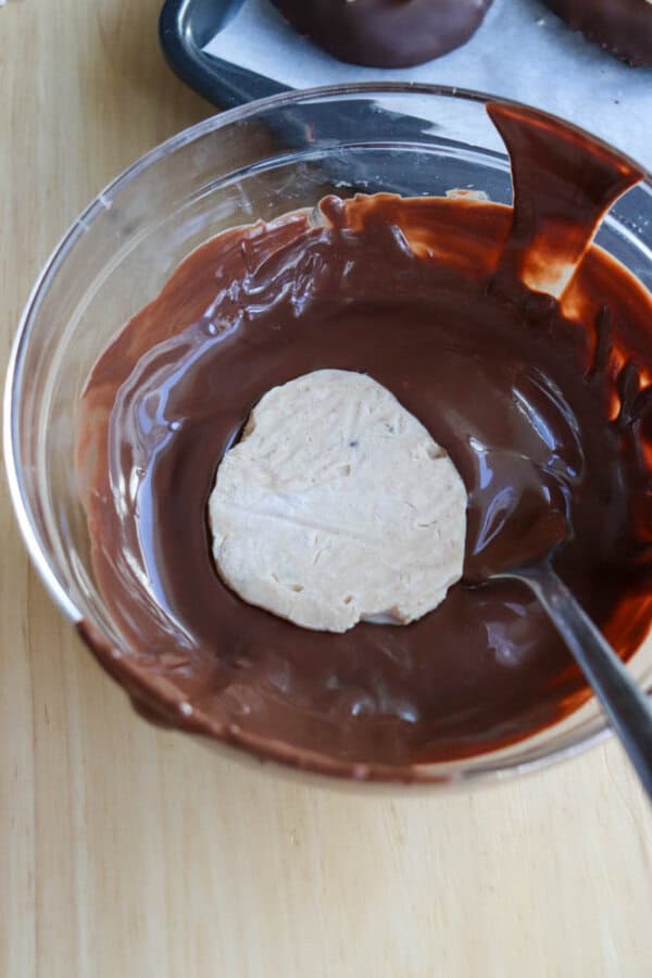 A bowl of melted chocolate with a round cookie partially dipped in it. A spoon rests inside the bowl, and a tray with more chocolate-coated cookies is visible in the background.