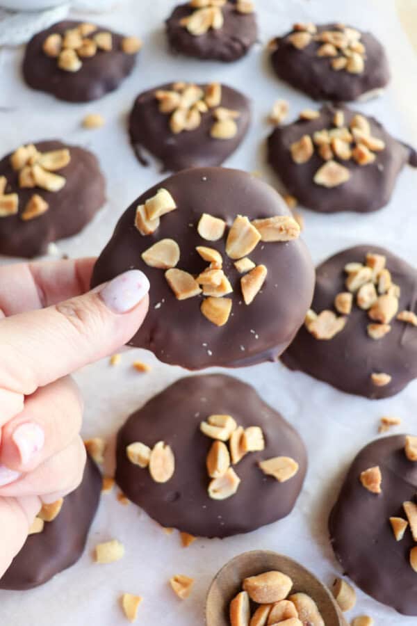 A hand holding a chocolate-covered cookie topped with chopped peanuts. More cookies with the same toppings are spread on a parchment-lined surface in the background. A small bowl of peanuts is partially visible.