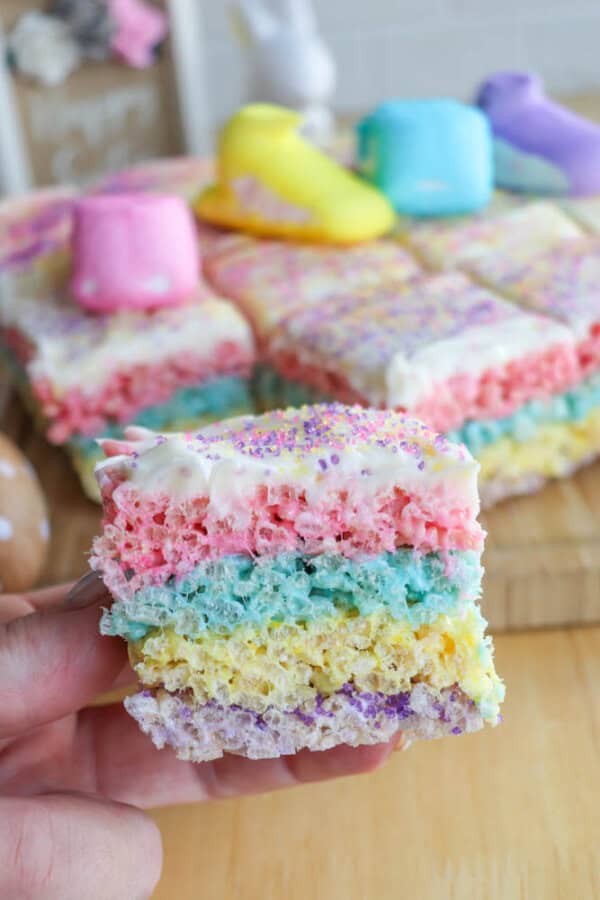 A hand holding a colorful, layered rice cereal treat with pink, blue, yellow, and purple layers topped with white icing and sprinkles. Blurred background shows similar treats and marshmallow shapes in pastel colors.