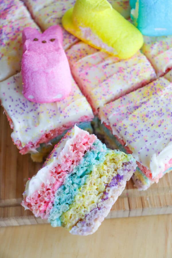 Colorful layered dessert squares with pastel pink, blue, yellow, and purple rice crispy layers, topped with white frosting and sprinkles. Decorated with pink, yellow, and blue chick marshmallows. Displayed on a wooden surface.