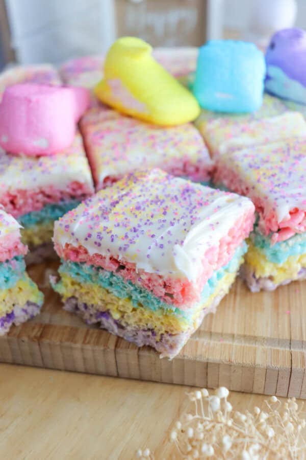 Colorful layered rice cereal treats topped with pink, purple, and yellow sprinkles sit on a wooden board. Marshmallow chicks in pastel colors are placed on top of some squares. The layers are vibrant in pink, blue, purple, and yellow.