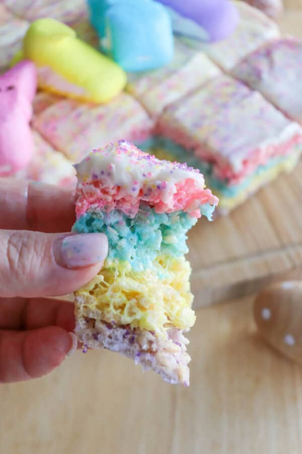 A persons hand holds a colorful, layered rice crispy treat, featuring pink, blue, green, and yellow sections. The treat is topped with white frosting and sprinkles. More treats are visible on a wooden board in the background.