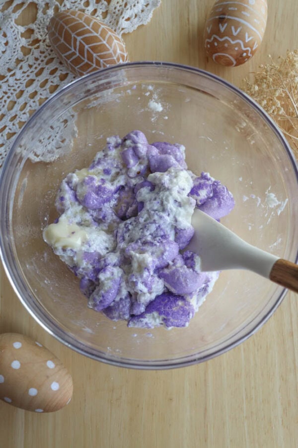A glass bowl filled with a mixture of purple and white dough, being mixed with a wooden spoon. The bowl is on a wooden surface, surrounded by decorative eggs and a lace tablecloth.