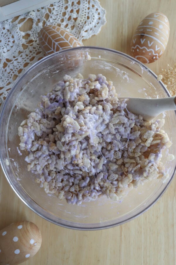A glass bowl filled with a mixture of cereal and purple marshmallow cream on a wooden table. A spatula rests inside the bowl. Decorated wooden eggs and a lace doily are nearby.