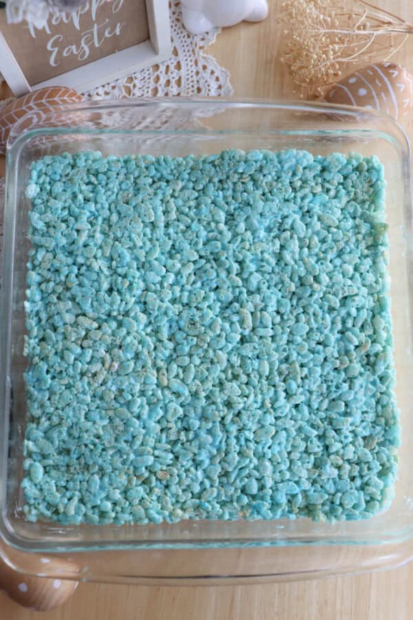 A square glass dish filled with blue rice cereal treats sits on a wooden surface. Decorative elements like dried flowers and a small Happy Easter frame are partially visible in the background.