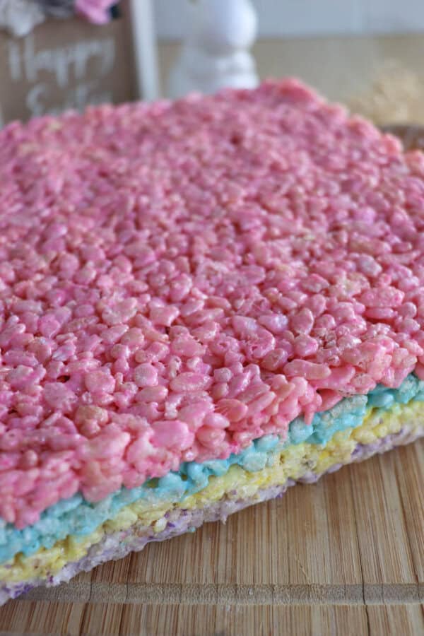 A close-up of multi-layered Rice Krispie treats with pastel-colored layers including pink, blue, yellow, and purple, displayed on a wooden surface.