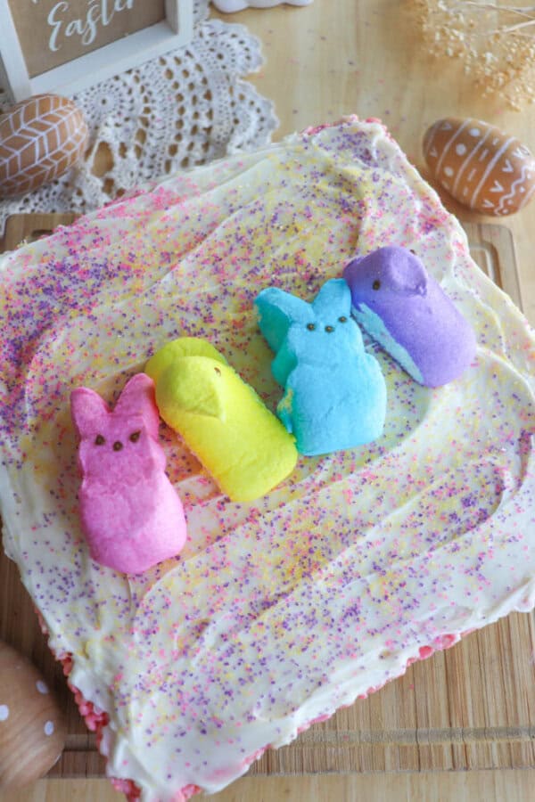 A square cake with white frosting is topped with colorful sprinkles and four marshmallow chicks in pink, yellow, blue, and purple. Wooden Easter egg decorations surround the cake on a wooden table.