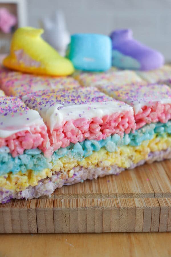 A colorful dessert made of layered rice cereal treats, featuring pink, blue, yellow, and purple layers. The top is sprinkled with purple and yellow sugar, and decorated with pastel marshmallow shapes. The cake is set on a wooden board.