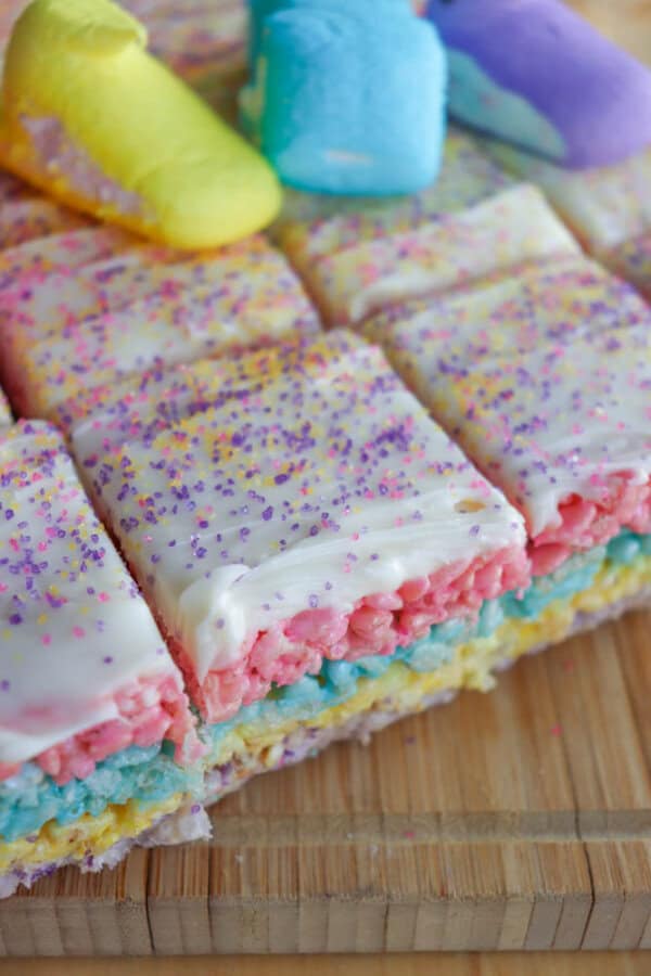Colorful layered Rice Krispies treats topped with white frosting and pastel sprinkles. Each layer is a different color: pink, blue, and yellow. Pastel marshmallows are scattered on top, all placed on a wooden board.