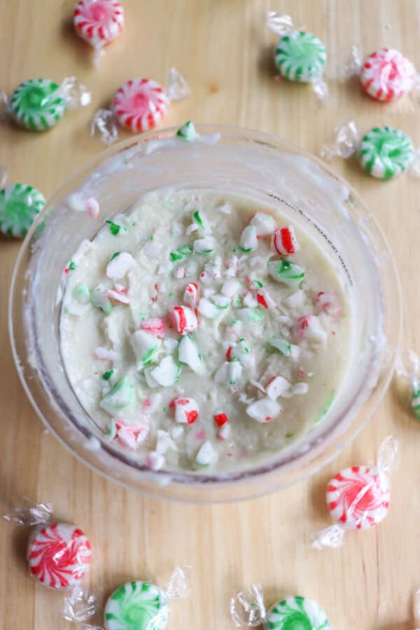 A bowl filled with creamy white ice cream topped with crushed red and green peppermint candies. The bowl is surrounded by whole red and green striped peppermint candies on a wooden surface.