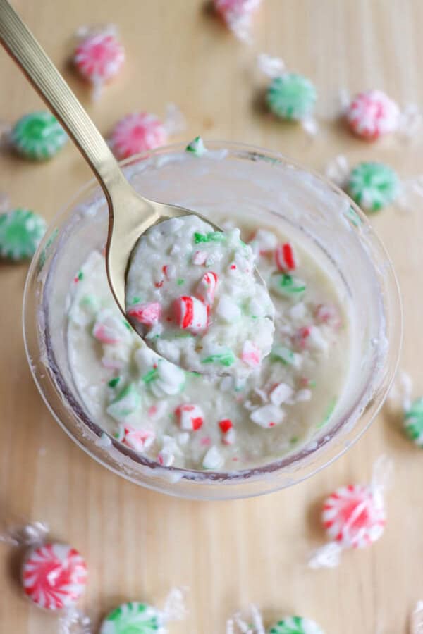 A bowl of white cake batter mixed with crushed red and green peppermint candies. A gold spoon scoops some of the batter. Whole wrapped peppermint candies are scattered on a wooden surface around the bowl.