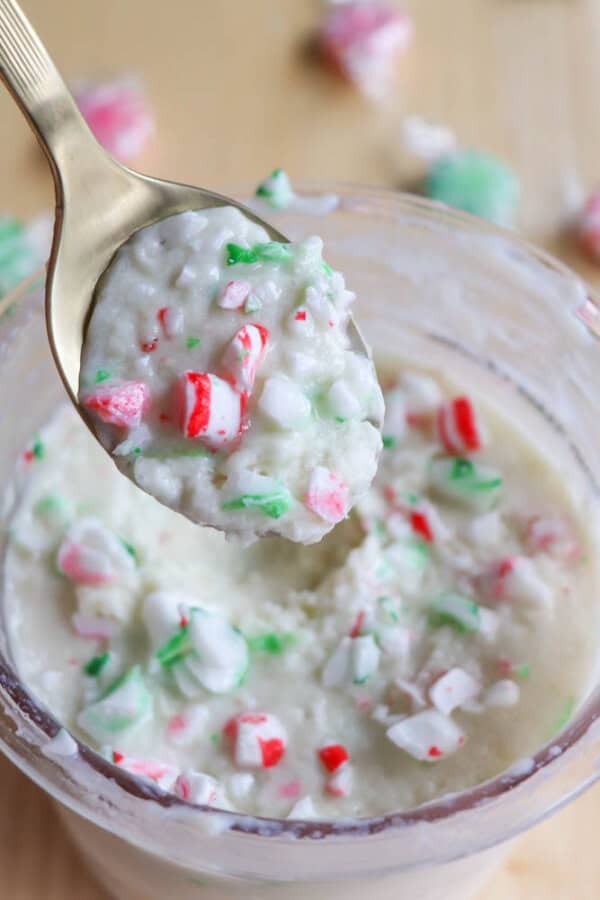 A spoon holds a creamy mixture with crushed red, green, and white peppermint candy pieces over a glass jar filled with the same mixture. Several unwrapped peppermint candies are scattered in the blurred background.