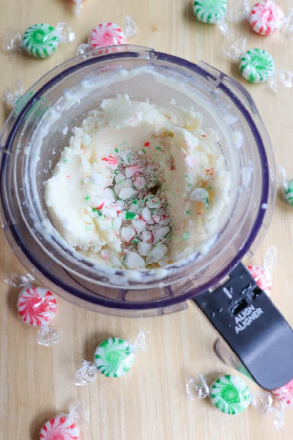 A food processor contains a creamy mixture with crushed red and green striped mints. Whole mints, some in clear wrappers, are scattered around the processor on a wooden surface.
