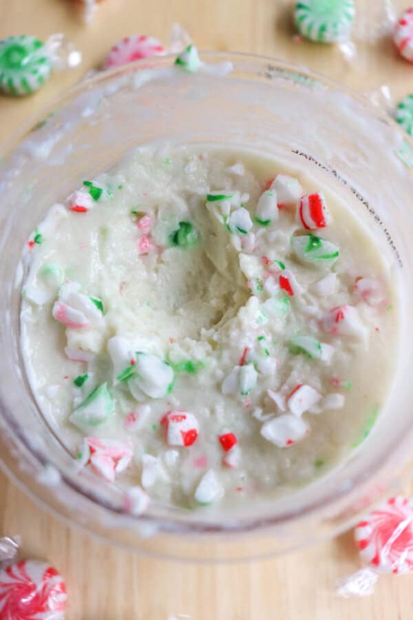 Close-up of a bowl containing a thick, creamy mixture topped with crushed red and green peppermint candies. Unwrapped peppermint candies are scattered on the wooden surface surrounding the bowl.