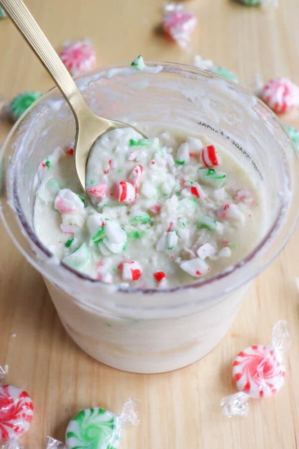 A cup filled with creamy white ice cream mixed with crushed red and green peppermint candies. A spoon rests inside the cup. Wrapped peppermint candies in red, green, and pink are scattered on a wooden surface around the cup.