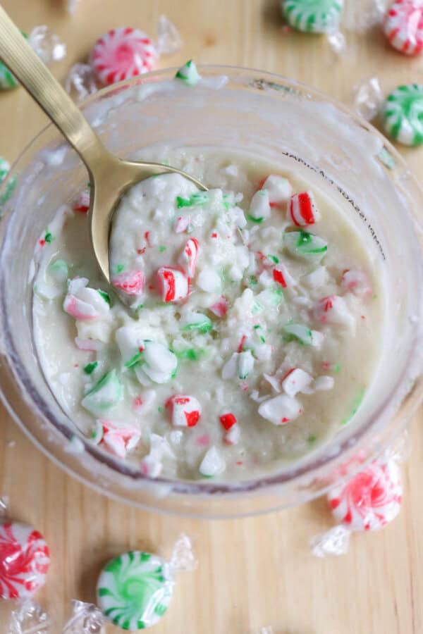 A bowl of peppermint sugar scrub with crushed red and green mint candies mixed in. A gold spoon is submerged in the mixture. Wrapped peppermint candies are scattered on the wooden surface around the bowl.