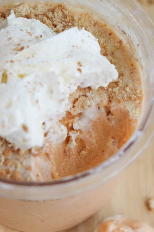 Close-up of a creamy pumpkin cheesecake ice cream with a swirl of whipped cream on top, sprinkled with crumbs. The caramel-colored delight, partially eaten, reveals a smooth texture beneath the topping. In the background, a soft, blurred wood surface adds warmth to this Ninja Creami recipe creation.