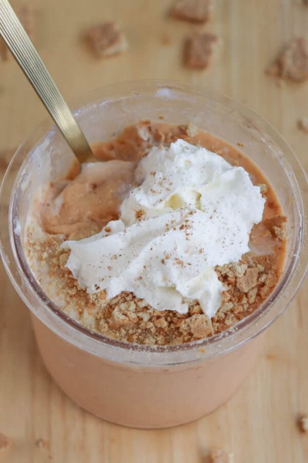 A creamy pumpkin protein ice cream in a clear cup topped with whipped cream, crumbled graham crackers, and a sprinkle of cinnamon. A golden spoon is sticking out of the dessert, resembling a raspberry cheesecake delight. Crushed crackers are scattered on the wooden surface around the cup.