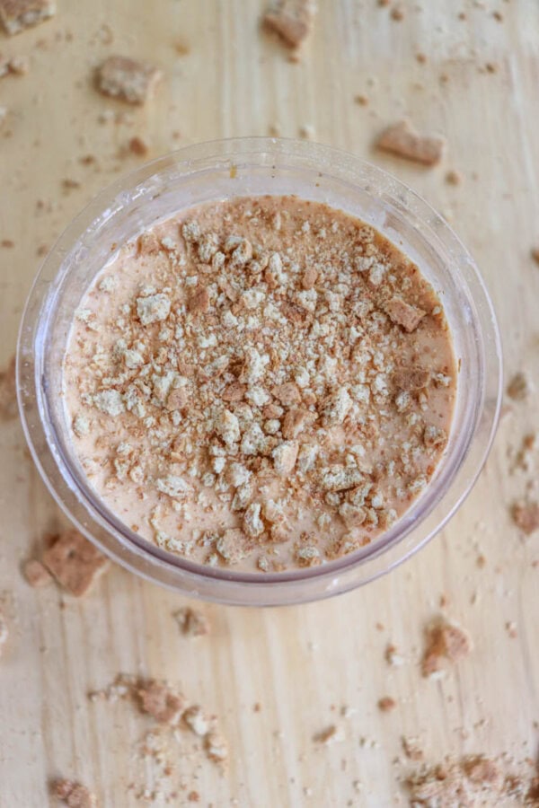 A glass bowl filled with a creamy, light brown dessert, reminiscent of a deconstructed raspberry cheesecake, sprinkled with crumbled cookies. More cookie crumbs are scattered across the wooden surface surrounding the bowl.