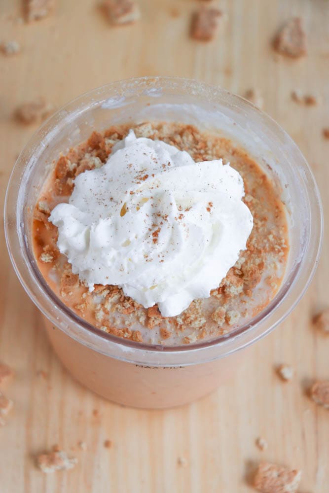 A plastic cup filled with creamy pumpkin protein ice cream, crowned with a swirl of whipped cream and sprinkled with crumbled cookies. Reminiscent of a ninja creami recipe, the cup sits on a wooden surface scattered with cookie crumbs.
