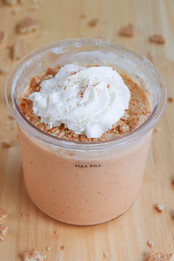 A creamy pumpkin ice cream in a clear pint, topped with whipped cream and sprinkled with crushed cookies. It sits on a wooden surface with crumbs scattered around.