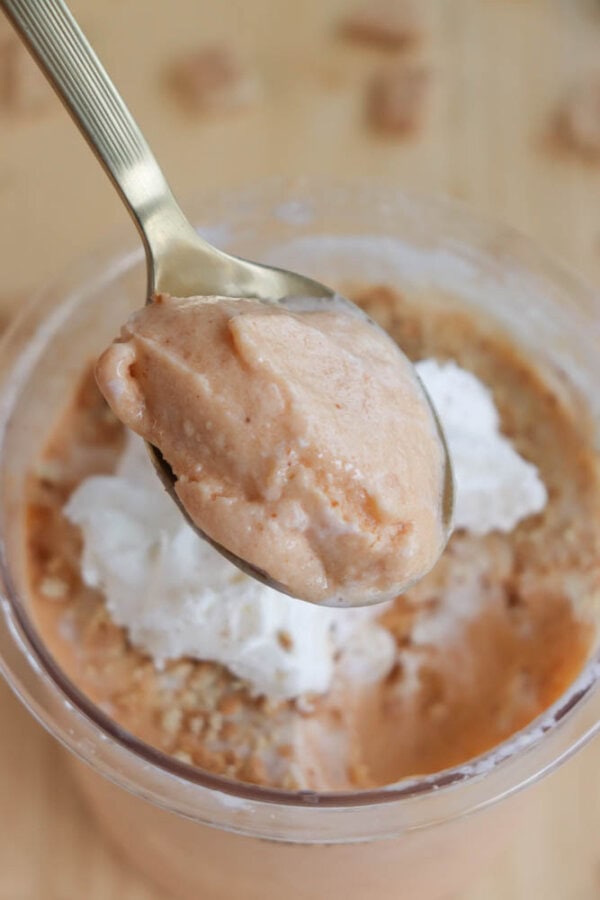 A spoonful of creamy, pumpkin protein ice cream is held above a bowl. The bowl contains the same ice cream, topped with dollops of whipped cream and crushed cookie bits on a wooden surface.
