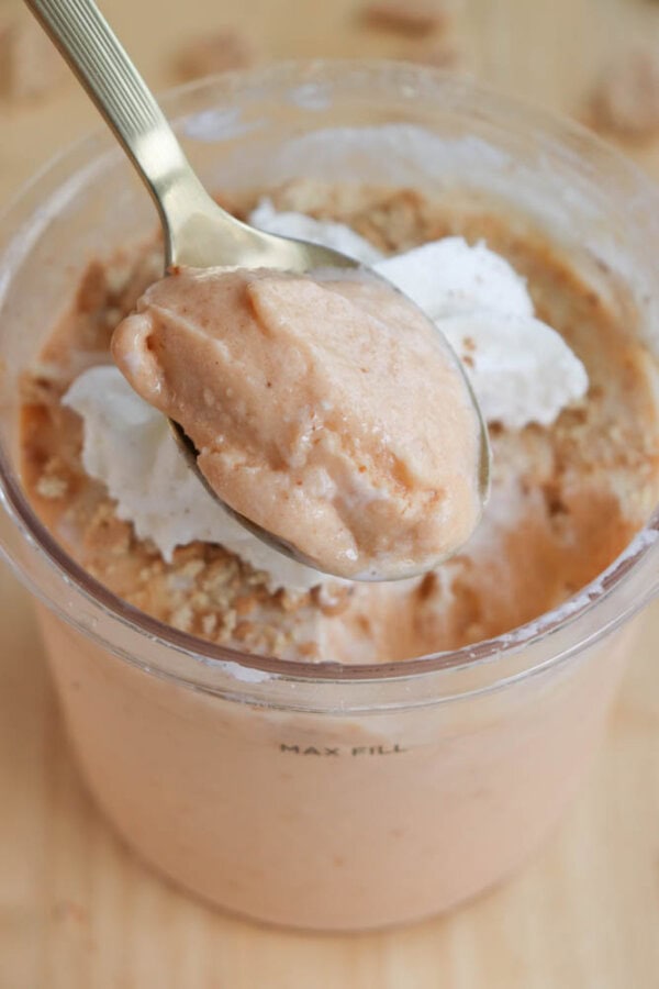 A close-up of a spoonful of creamy, light orange dessert being lifted from a cup evokes the delightful sensation of a ninja creami recipe. Topped with fluffy whipped cream and sprinkled with crumbs, the silver spoon contrasts beautifully against the MAX FILL marked cup.