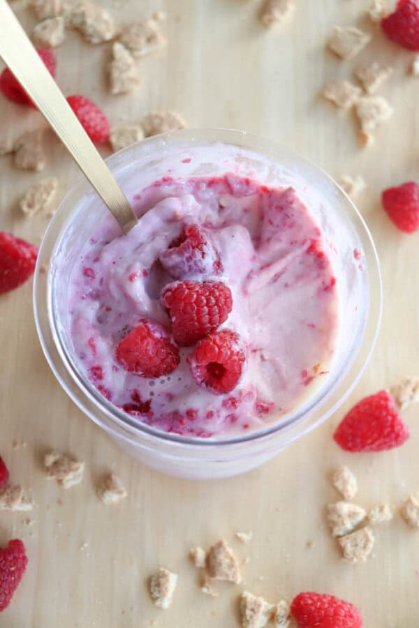 A bowl of raspberry yogurt with a gold spoon, topped with fresh raspberries. Crumbled cookie pieces and whole raspberries evoke the rich flavors of blueberry cheesecake ice cream, scattered across a rustic wooden surface.