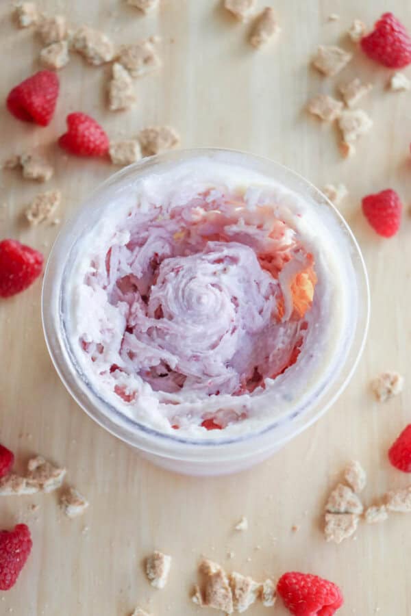 A swirl of raspberry-flavored ice cream in a clear container on a wooden surface, surrounded by scattered graham cracker crumbs, fresh raspberries, and hints of blueberry cheesecake ice cream for an indulgent twist.