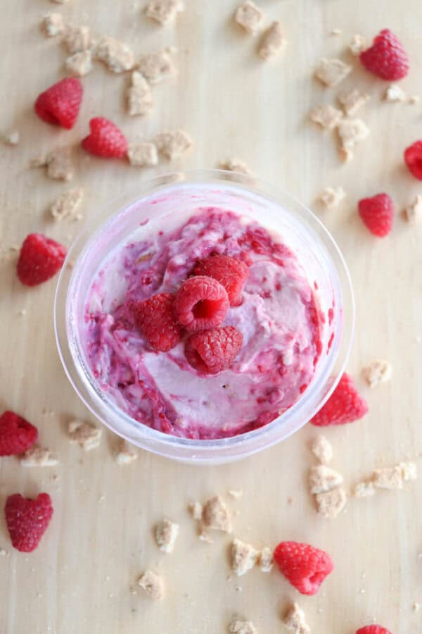 A cup of raspberry mousse topped with fresh raspberries, surrounded by scattered raspberries and crumbled pieces of dessert on a wooden surface, creates a scene reminiscent of blueberry cheesecake ice cream dreams.