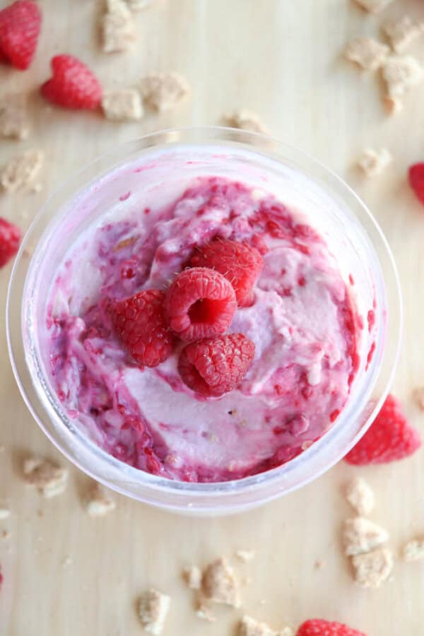 A bowl of pink raspberry ice cream topped with fresh raspberries, reminiscent of blueberry cheesecake ice cream. Crumbled pieces, possibly granola or cookies, are artfully scattered around the bowl on a light wooden surface.