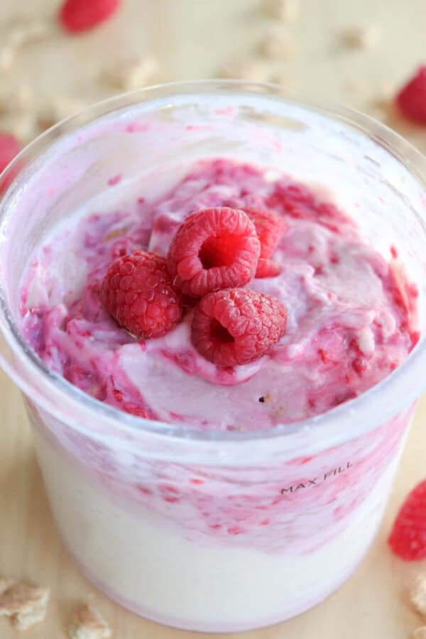 A clear container filled with swirled ice cream and raspberries resembles a creamy mix of white and pink, like blueberry cheesecake ice cream. Several fresh raspberries are placed on top as garnish, while crumbled pieces and whole berries rest in the background.