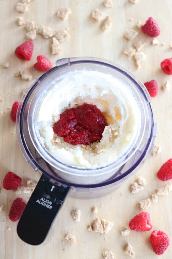 A food processor contains a creamy white mixture reminiscent of raspberry cheesecake ice cream, topped with red raspberry sauce and crumbled graham crackers. Raspberries and more crumbled graham crackers are scattered on the wooden surface around the processor.