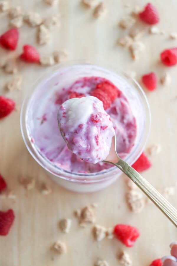 A spoonful of raspberry ice cream is held above a jar brimming with creamy goodness, reminiscent of blueberry cheesecake ice cream. Raspberries and small granola pieces are artfully scattered on a light wooden surface.