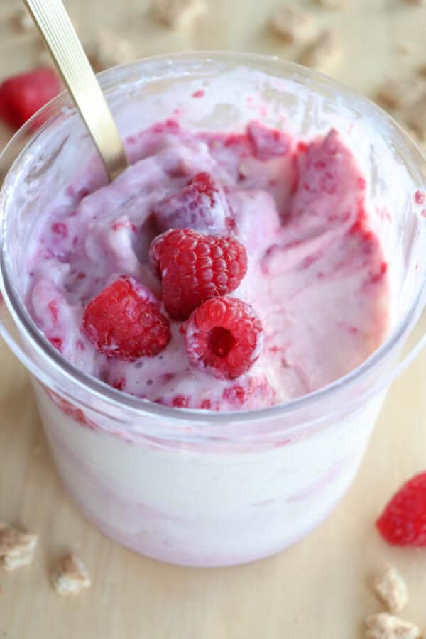 A glass cup filled with creamy ice cream mixed with raspberries evokes the indulgent delight of blueberry cheesecake ice cream. Several whole raspberries garnish the top, while a spoon is partially dipped into the smooth mixture, surrounded by small crumbly pieces.