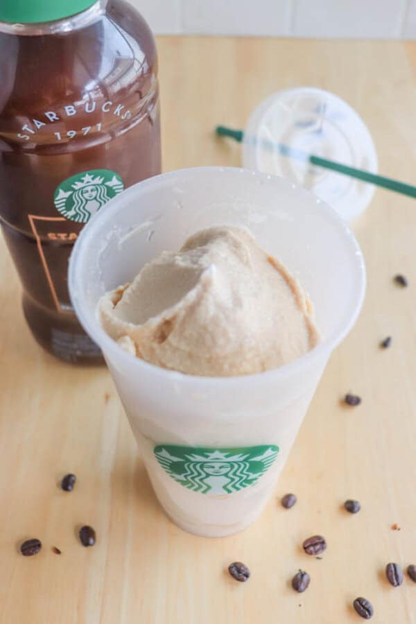 A Starbucks cup filled with a blended coffee drink sits on a wooden table. Coffee beans are scattered around, and a bottle labeled Starbucks is partially visible in the background next to a plastic lid with a straw.
