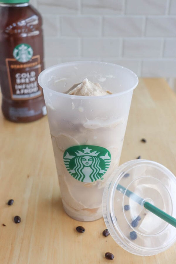 A cup filled with a blended beverage, featuring the Starbucks logo, sits on a wooden table. A clear lid and green straw lie next to it. Coffee beans are scattered around. A Starbucks cold brew coffee bottle is in the background.