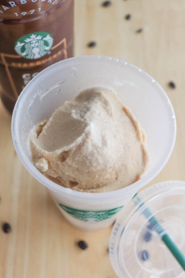 A plastic cup containing light brown ice cream sits on a wooden table. The cup has the Starbucks logo. There is a drink bottle and a lid with a straw nearby, and a few coffee beans are scattered around.