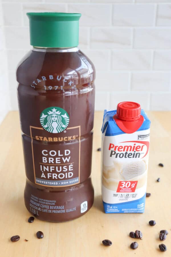 A large bottle of Starbucks cold brew coffee is next to a small carton of Premier Protein vanilla shake on a wooden surface. Coffee beans are scattered around them. The background is a white-tiled wall.
