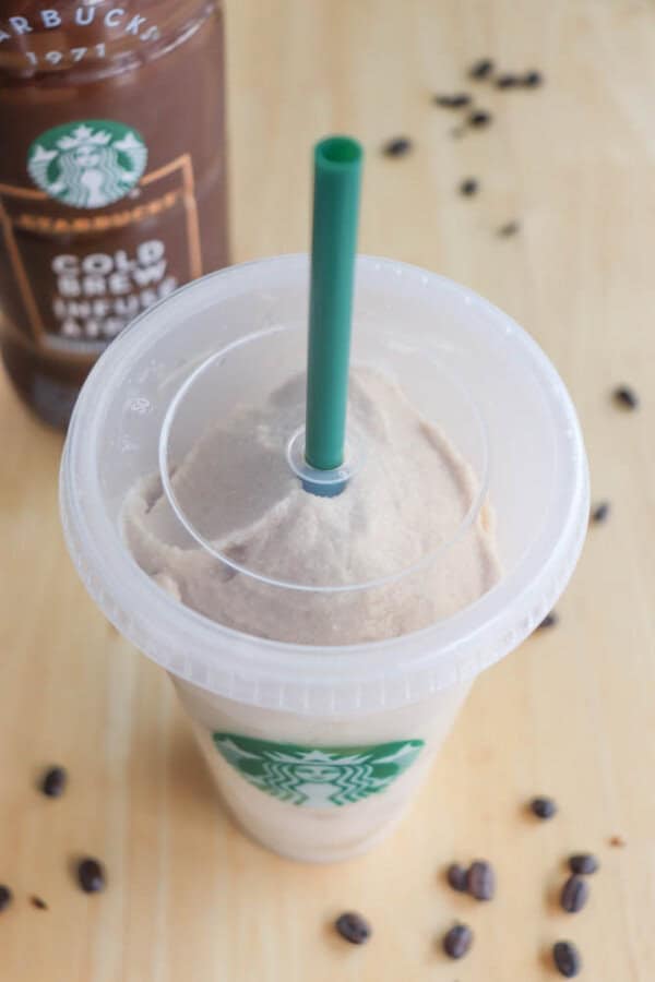 A Starbucks cold brew coffee in a clear plastic cup with a green straw. The drink has a creamy foam on top. In the background, there is a bottle of Starbucks Cold Brew Coffee and scattered coffee beans on a wooden surface.